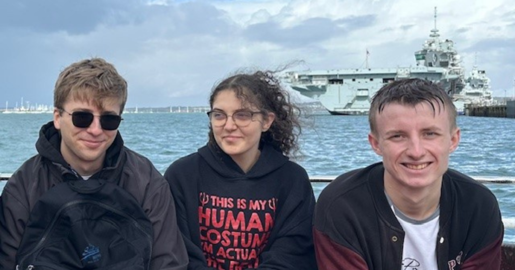 Three students sat on boat with an aircraft carrier in the background